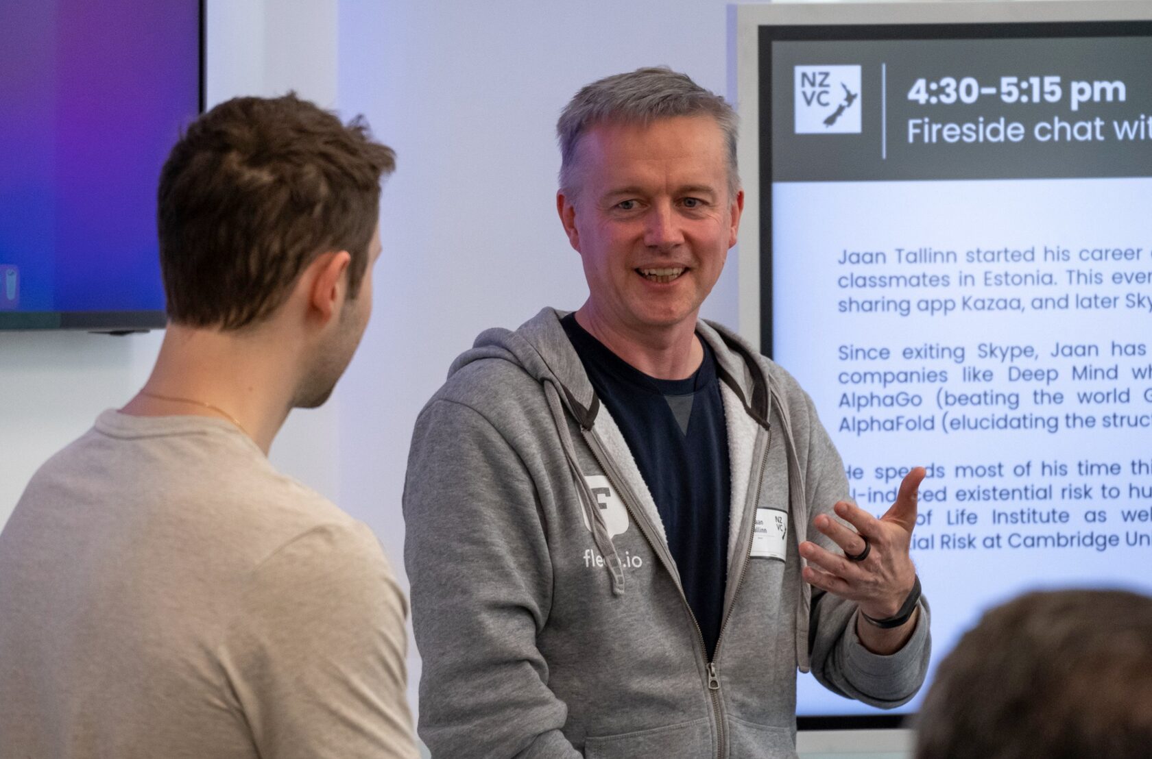 Two attendees talking beside the screen billing the fireside chat with Jaan Tallinn