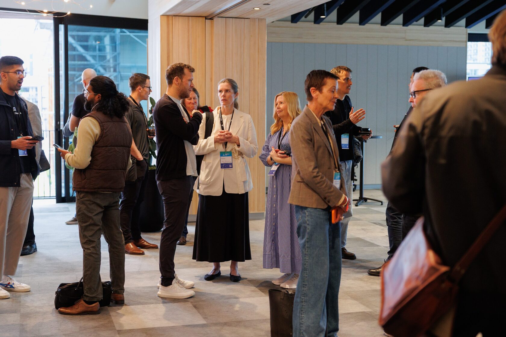 A dozen attendees standing with coffee cups and lanyards in the timber-lined Pipiri Lane foyer