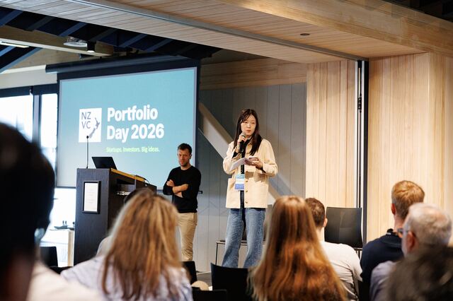 A host opening the day on stage beside a screen reading NZVC Portfolio Day 2026